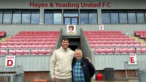 A tall man with dark hair and a shorter man with silver hair embrace and smile at the camera in front of red seats in the stands of a football stadium