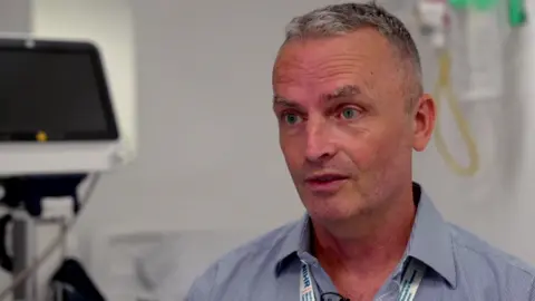 A doctor with short grey hair wears a blue shirt and a white lanyard. He is stood in a hospital room with medical equipment behind him.