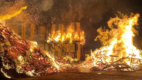 Taunton Fire Station A large fire in Somerset. There are separate piles of wood and wood chip alight, with big orange and yellow flames.