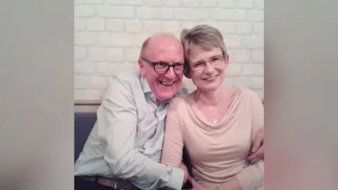 A man and woman are sat on a bench with their arms interlinked. The man on the left is wearing a pale blue shirt and glasses, the woman on the right is wearing a pale pink shirt and glasses.