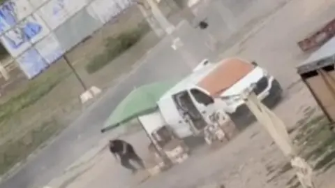 Man dressed in black chased by a black drone as he runs close to a white van and a green parasol on a dusty bit of ground alongside a road.