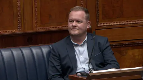 A man sits in front of a mic. He's wearing a suit. Wooden panelling is on the walls behind him.