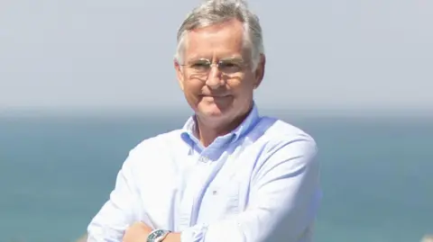 St Moritz Hotel and Cowshed Spa A close up of Hugh Ridgway. He has short grey hair, is wearing a white shirt. In the background is a huge body of water.