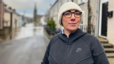 A woman, wearing glasses, a cream hat and a black Nike zip up hoodie. She is standing in front of a flooded street, which is blurred in the background.
