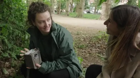 Two people crouching beside a fence in a park, one holding a small wildlife camera device among leaves.