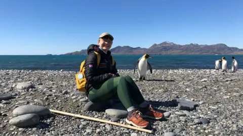 Aoife McKenna Aoife McKenna in Antarctica. She is wearing a cap and glasses, and sitting down on a rock by the shore, with four penguins and the ocean behind her 