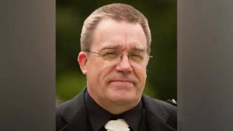 Police Scotland A man with short brown hair wearing a dark suit, white tie and glasses.