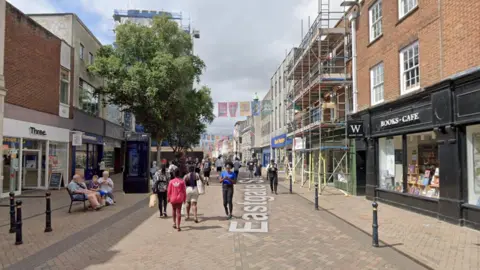 Google maps street view of Eastgate Street in Gloucester. There are shops and businesses lining both sides of the street and there are flags hanging above the street. Lots of people can be seen walking around in the area.