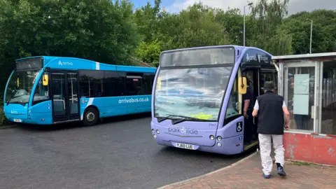 A blue bus and purple bus parked side by side next to a bus stop and a man wearing a blue gilet and hat about to board