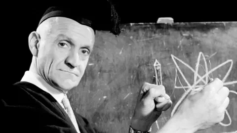 Stanley Unwin wearing a black academic cap and a suit. He is standing in front of a chalkboard.