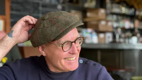 A smiling man with round tortoiseshell glasses and a tweed flat cap with a tattoo on his wrist sitting in a pub