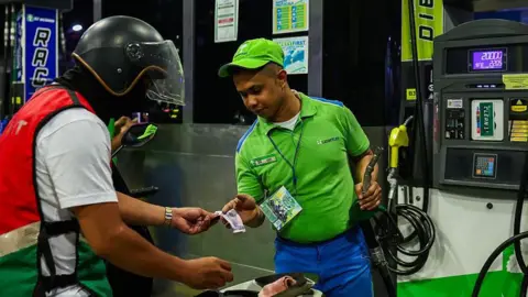 A worker in a green shirt and cap collects payment from a motorcyclist after refilling his motorcycle with petrol at a gas station in the Philippines