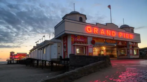 Weather Watchers/Gary the monkey Weston-super-Mare pier is pictured at sundown. At the entrance the words 'Grand Pier' stand out in almost a neon orange/red colour. That light is reflected on the cobbles in front of the entrance. The beach and sea can also be seen and there are dark clouds passing overhead. 