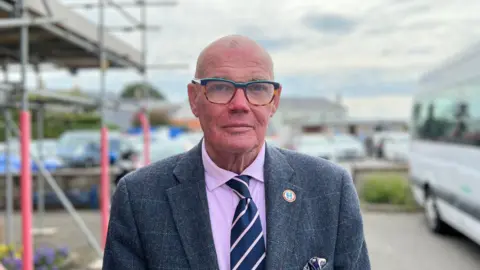 Constable Marcus Troy. He is bald and wears glasses. Mr Troy is wearing a blue wool suit with a subtle check. He is also wearing a purple shirt with a navy blue, purple and white striped tie. The background is blurred but appears to be a car park with a small bus to the right of the picture.