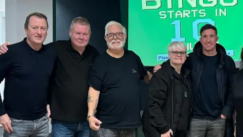 TONY GRANT Tony Grant and Robbie Fowler stands alongside several people at the centre. They're dressed in dark clothes and smiling - with their arms around each others shoulders.