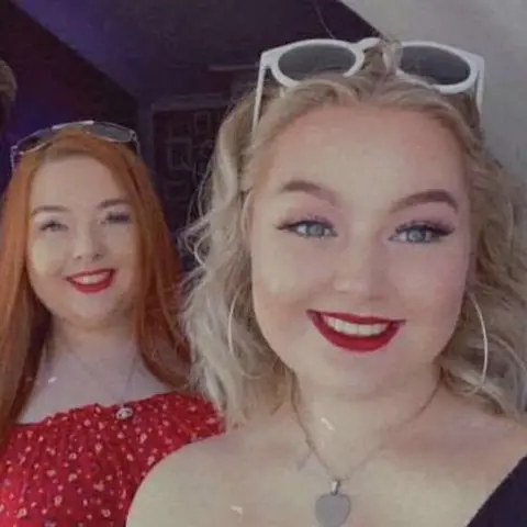 Jessica Thomas Two girls with sunglasses on their heads and red lipstick, smiling at the camera. One has long straight red hair, the other short curly blonde hair. The one with red hair is stood behind the other and has a red top on. They both silver have necklaces with pendants on. 