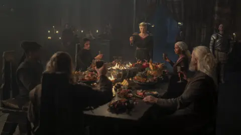 HBO The House of the Dragon group, sitting around a sumptuous feast, raise their glasses to toast