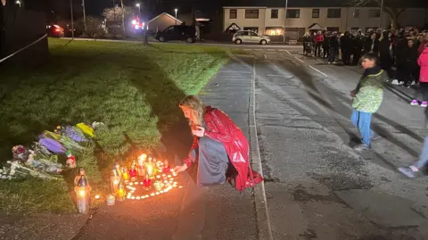 A woman is crouching on the ground to lay a candle on a memorial on a street. Candles are lit and flowers are lying on the grass. A ground of people are watching from across the street.