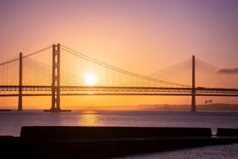 Amy Jenkins The sun sets over the Forth Road bridge from South Queensferry. The sky is orange and the water is sparkling and dark purple.