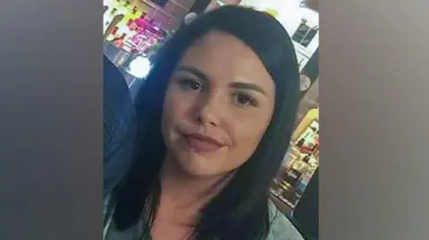 Alice Webb is smiling at the camera, she has dark hair and a piercing in her lower lip. She stands in front of an illuminated bar.