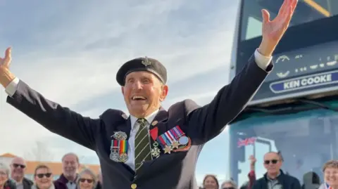 An old man wearing a military uniform and medals stands smiling with his arms outstretched in front of a bus. People are standing and smiling in the background. 