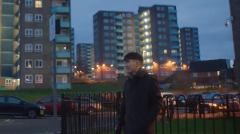 Tell Studio/Hero Images A man in his twenties wearing a dark coat stands on a street corner at dusk and appears pensive. Tower blocks and parked cars are behind him.