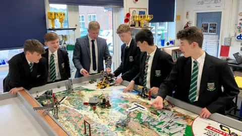 Five pupils wearing school uniforms, including dark blazers and green ties, and a man wearing a suit gather around a table in a large science classroom. The table has a big map on it with Lego structures in various places.