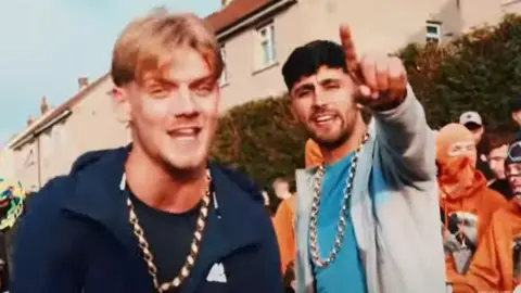 Two young men wearing chains pose on a residential street and point at the camera