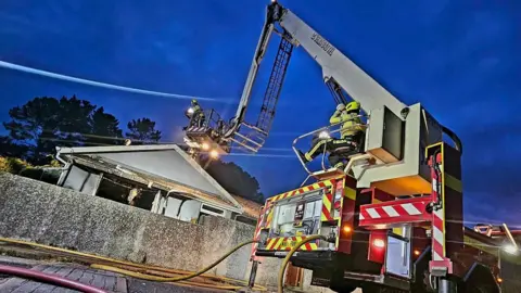 Devon and Somerset Fire and Rescue Service / Buckfastleigh Fire Station An aerial ladder platform carrying a firefighter who is helping tackle a blaze at a bungalow