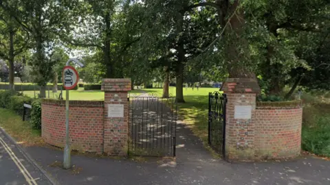 Google A corner entrance to the park in North Walsham, featuring two sections of curved red brick wall either side of a black wrought iron gate. Beyond the gate, a concrete path goes off into the distance, lined by tall trees with lawns either side.