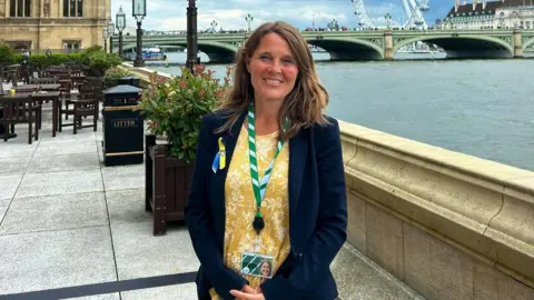 Vikki Slade Vikki Slade stands outside with the London Eye in the background and a bridge over the River Thames. She is smiling at the camera, has mid length light brown hair and wears a navy blue blazer with a yellow and white floral print top underneath. 