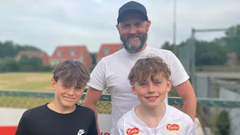 Owen Sennitt/BBC A man wearing a hat and white T-shirt smiles towards the camera alongside two boys with houses blurred in the background