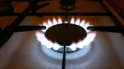 A close-up of a lit gas ring on a white hob. The flames coming out of the dark ring are bright white with red tips.
