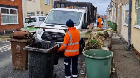 North East Lincolnshire Council A council worker in a high vis jacket and work trousers with stripes is loading up bins onto a council truck. The vehicle is parked on a street of terraced houses. One resident is watching from his doorway.