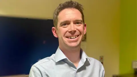 SUPPLIED Man in blue short with short brown hair smiling infront of yellow wall and window with blue blinds 