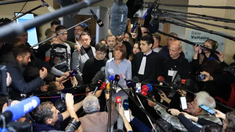 EPA Gisèle Pelicot is framed in the midst of a wide dramatic dramatic shot inside the court, with a large crowd of journalists surrounding it with microphones