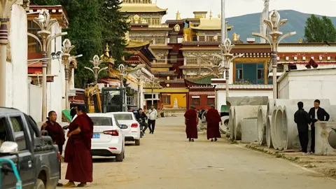 Xiqing Wang/ BBC Monks and other on a road leading to the Kirti monastery. Parked cars flank the road.