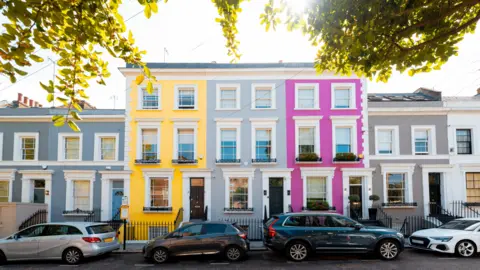 Getty Images A well-to-do row of terraced houses in Notting Hill with high end cars parked in the street. The houses are painted in vibrant colours. 

