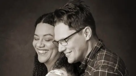 A black and white image of David Clark, wearing glasses and a plaid shirt, embracing his wife.