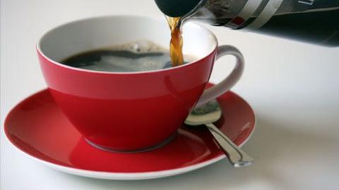 A red coffee cup with a spoon sitting on top of a saucer.  A cafitiere is seen pouring coffee into the cup