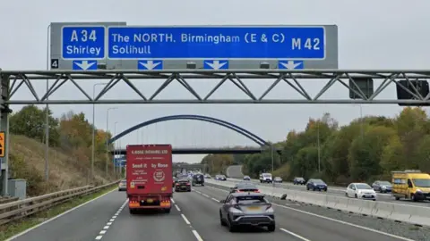 A motorway with cars and lorries on it and a blue sign overhead with Birmingham, Solihull and Shirley written on it 
