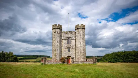 A tower stands in the middle of a field with one door, laying out on a field. Clouds can be seen in the background.