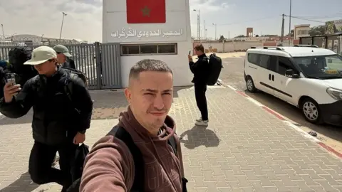 @rucktravels Tom Ruck taking a selfie successful  beforehand   of the Guerguerat borderline  crossing betwixt  Western Sahara and Mauritania. Behind him is simply a ample  factual  operation   with a Moroccan emblem  painted connected  it.