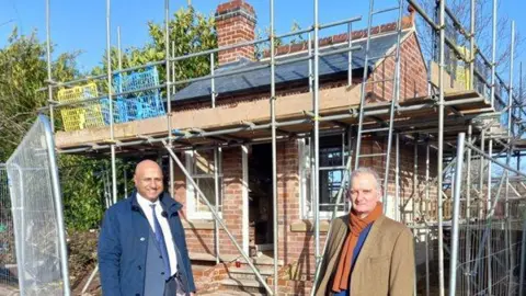 Two men stand outside a small one-storey redbrick building, which is one of the 19th century summer houses. The man on the left, councillor Naeem Akhtar, smiles while wearing a grey suit and blue coat. The man on the right, Nicholas Dutton from Historic England, wears an orange scarf, blue shirt, and brown jacket. The summer house is covered in scaffolding.