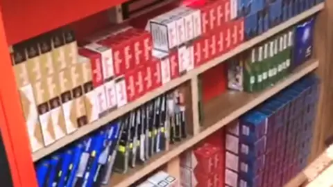 Essex Trading Standards Cigarette boxes and tobacco pouches stashed under a shop counter, stored on wooden shelves.