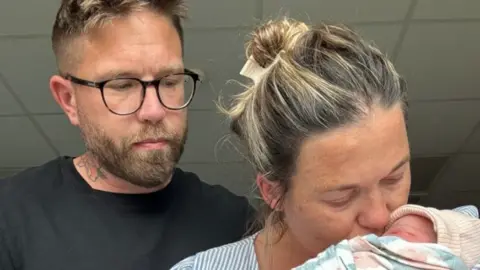Aaron Way A man looking on as a woman cradles a baby swaddled in a pink and blue muslin, with a striped cap. The woman kisses the baby's face. He is in a black t-shirt, wearing black glasses and has spiked brown and blond hair. The woman has blond hair clipped back and a white-and-blue striped top.