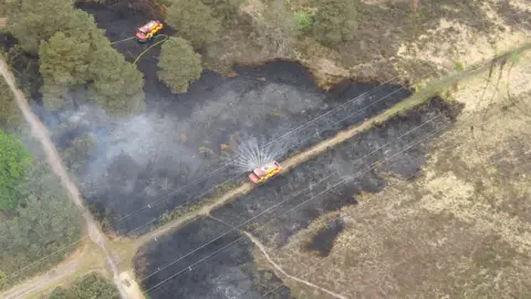 Surrey Fire and Rescue Service An aerial view of the fire being put out by two engines which are parked on the land and on a path. There is water spraying from the side of one engine and the plants on the ground are black with smoke rising up