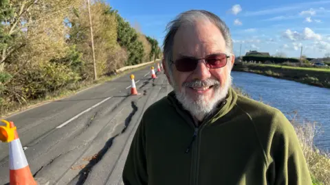 Alan Whitney, 76, has a grey beard and moustache. His black and grey hair is being blown across his head by the wind. He has glasses on with a tint on the lenses and he is wearing a bottle green fleece. Behind is a badly cracked road with orange and white cones near the middle white lines. On the right is a waterway.