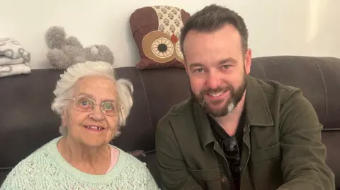 SDLP An elderly woman to the left with grey hair and glasses. She is wearing a light green jumper. To the right is a bearded man who is wearing a green jacket with a black top and sunglasses hanging from it. They are sitting on a brown sofa with to cushions and a grey teddy bear sitting on top.