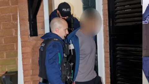 Police officers lead a man out of a house into a police van